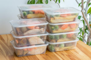 Three individual stacks of clear plastic containers filled with prepared meals. Each stack is 3 or 4 containers high that include colorful orange and green vegetables that are sitting on a butcher block table. 