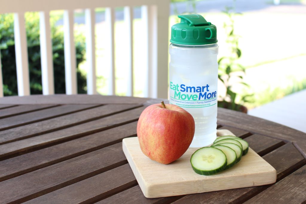 An apple, cucumber slices, and a clear water bottle sit on top of a wooden board outside.